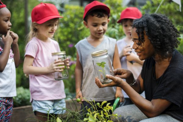 A woman shows children something in a jar. They are all in a garden.