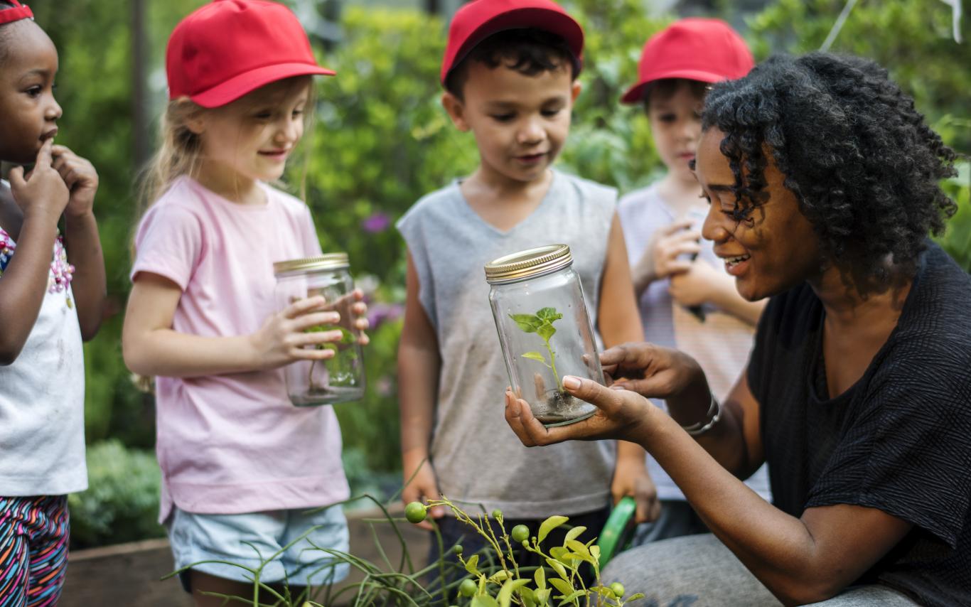 A woman shows children something in a jar. They are all in a garden.