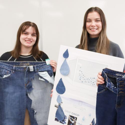 One students holds a pair of jeans with fabric cut out of it and another holds a poster.