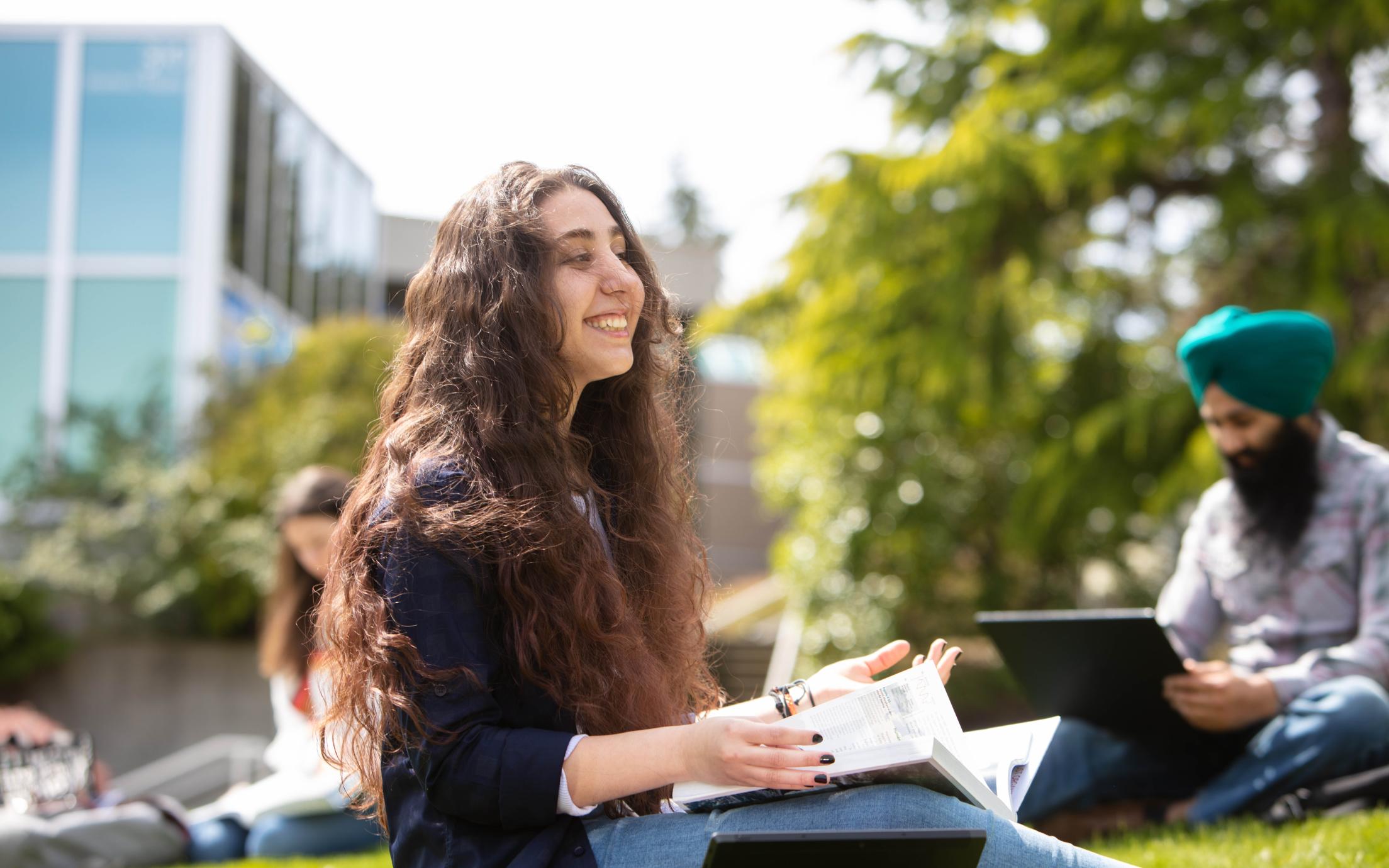 Student sits on grass with her books open with other students in the background