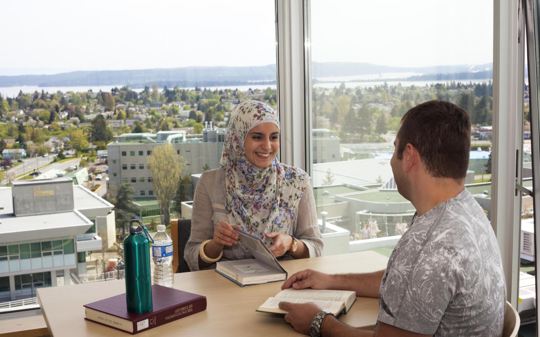 man and woman smiling while sitting across from one another at a table by a window. textbooks are on the desk in front of them