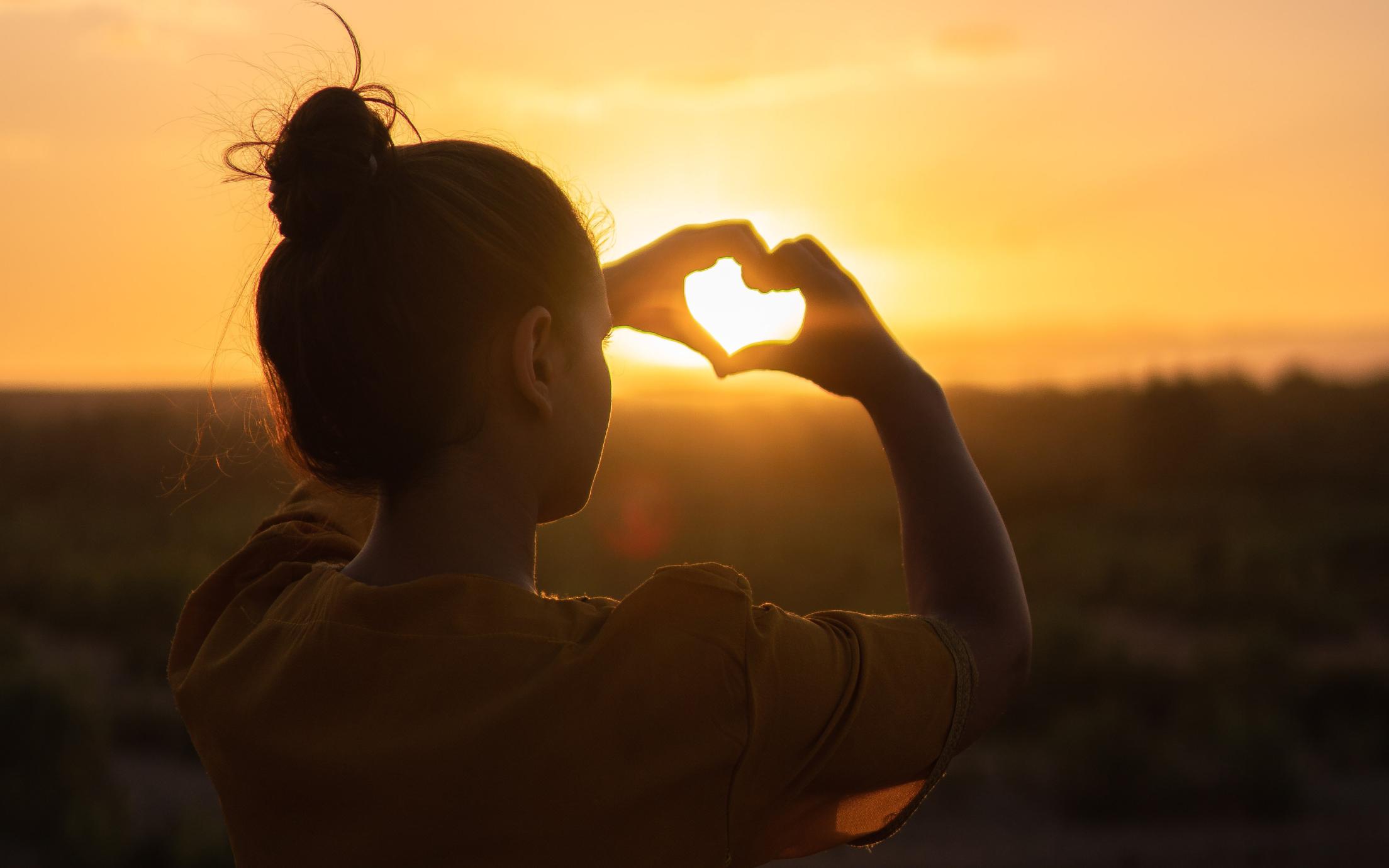 Girl making a heart with her fingers and the sunset poking through