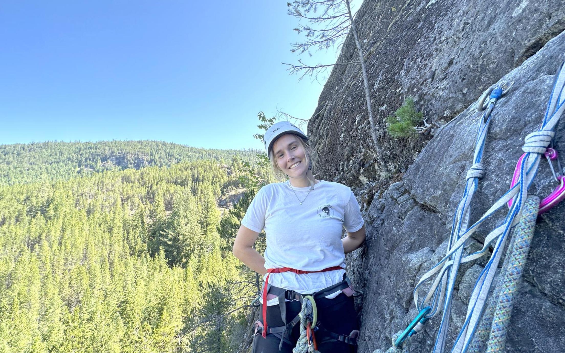 Sydney Solland stands on a ledge next to a rock face with climbing gear in front of her