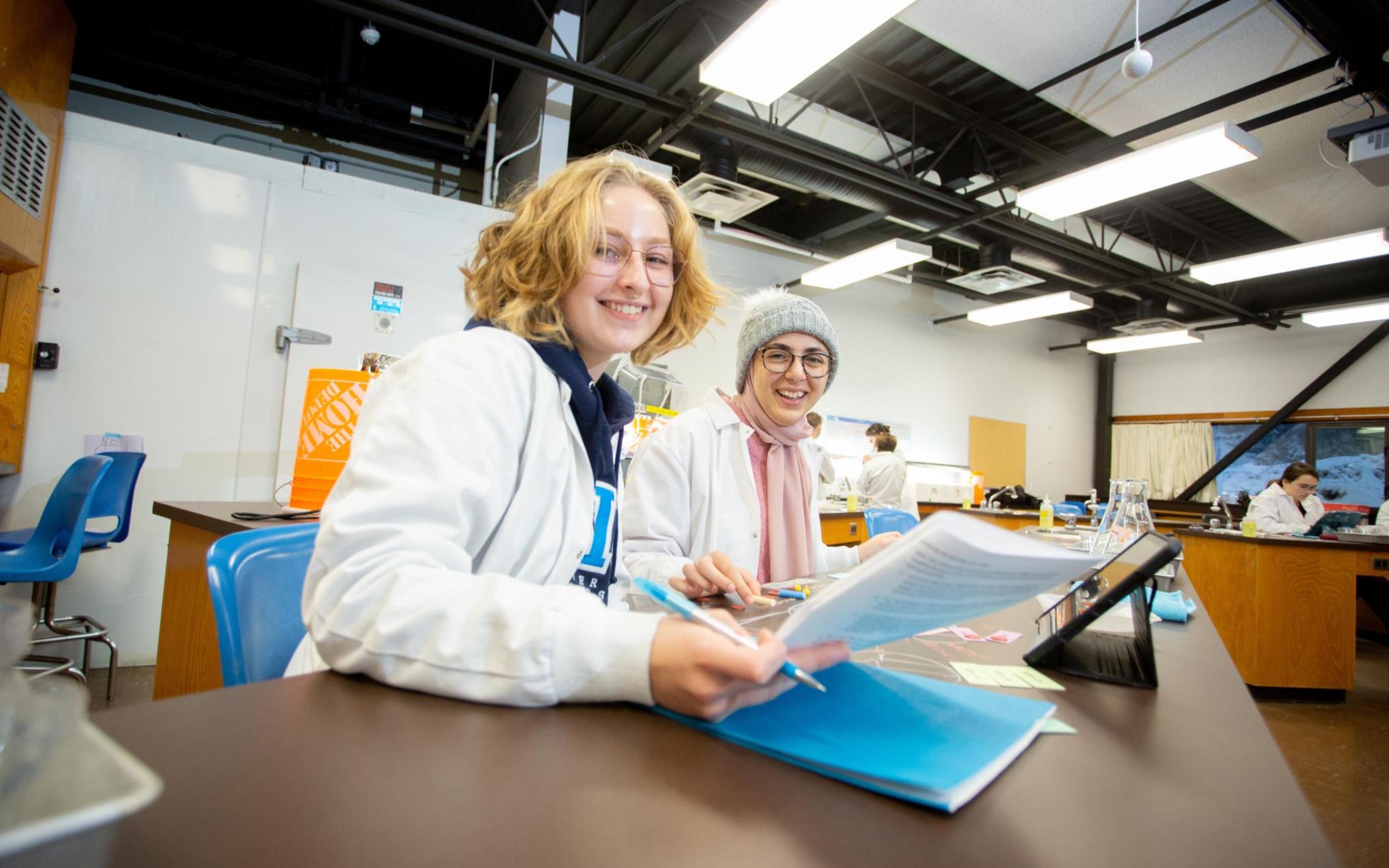 Two students look at a sheet in a lab, one has a pen in her hand, they are both smiling at the camera