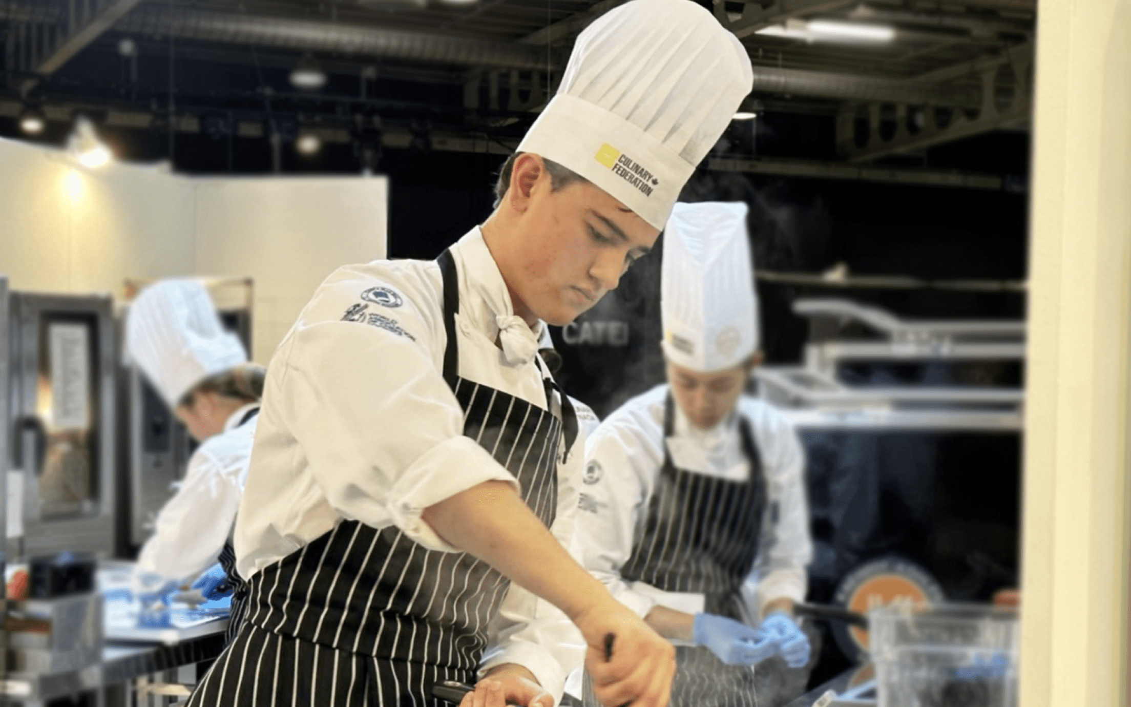 Ottis Crabbe wearing chef's hat and full cooking attire preparing a dish in a pot on a stove in an industrial kitchen during a competition.