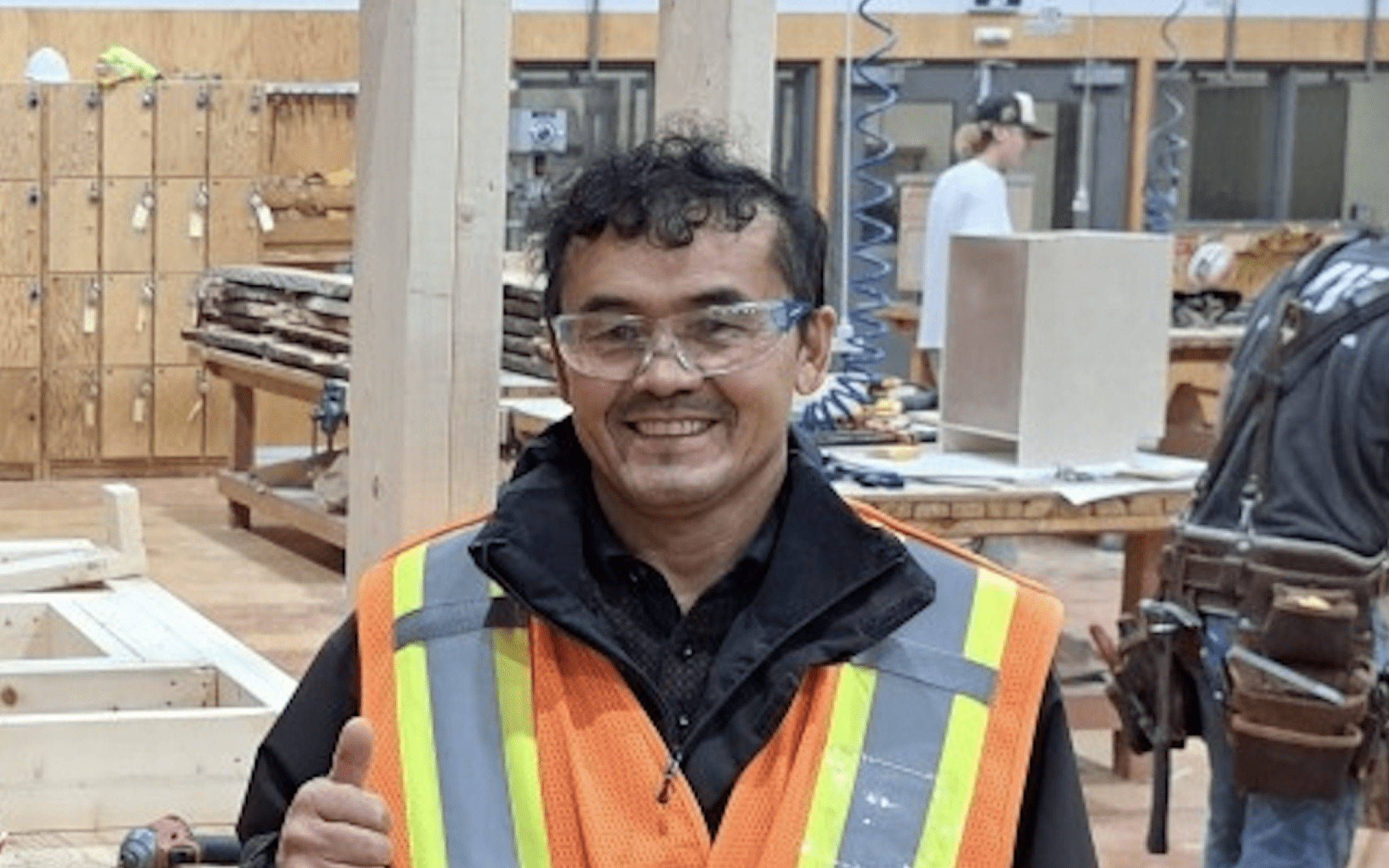 Ezat Haidary wering a reflective vests and safety goggles in the carpentry shop, giving a thumbs up and smiling at the camera.