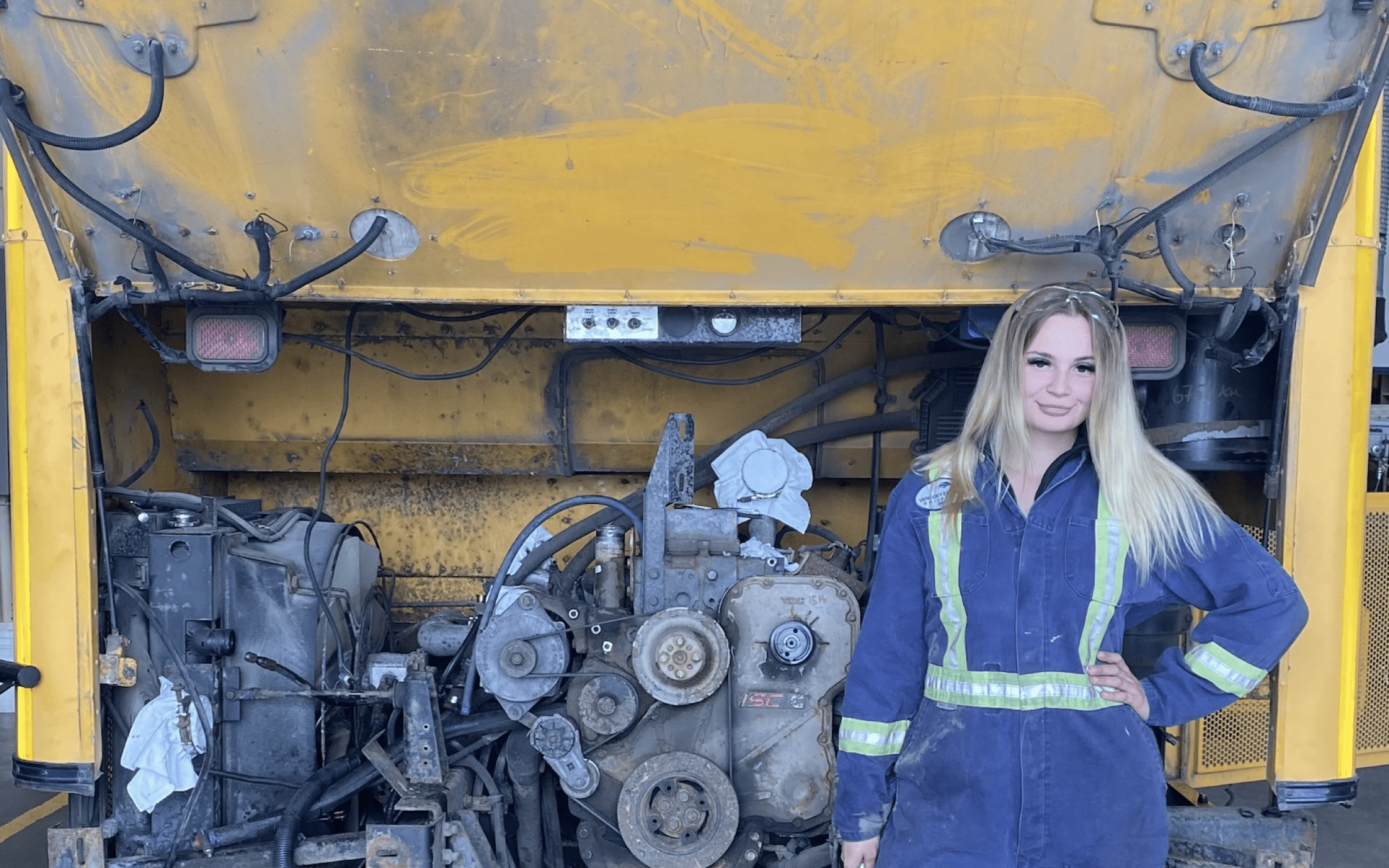 Madeline kozubal wearing mechanics coveralls standing in front of a school bus engine and smiling at the camera