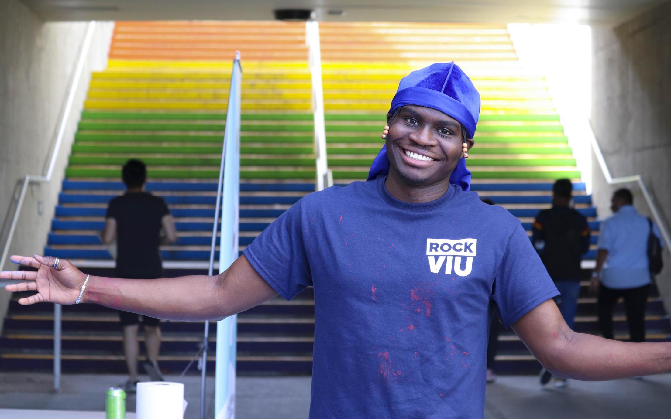 Black student holds arms out in welcome standing in front of the rainbow stairs