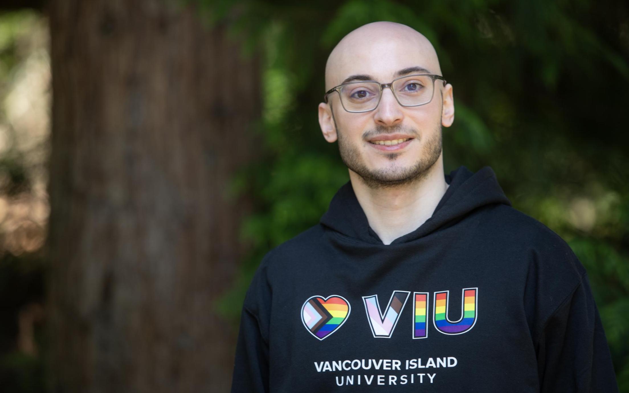 VIU grad and current employee Rayan Zeineddine wearing a VIU hoodie and glasses standing next to a tree outside and smiling at the camera
