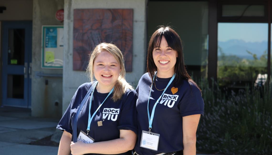 Two women wearing RockVIU shirts and lanyards smile in front of Building 310 at VIU's Nanaimo campus
