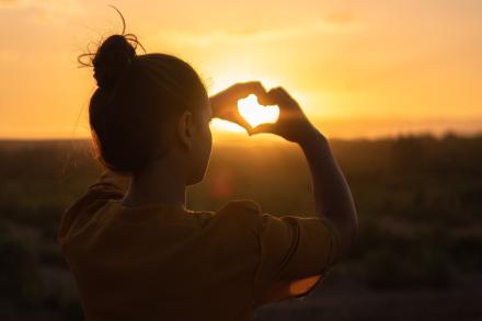 Girl making a heart with her fingers and the sunset poking through