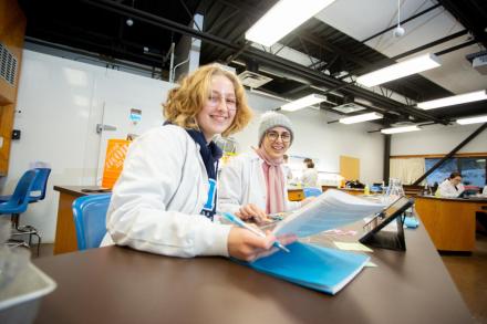 Two students look at a sheet in a lab, one has a pen in her hand, they are both smiling at the camera