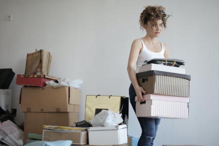 Student carrying boxes with more piled behind her, looking anxious