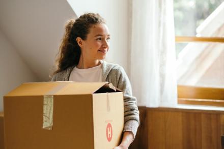 A girl holds a moving box while standing in a bedroom