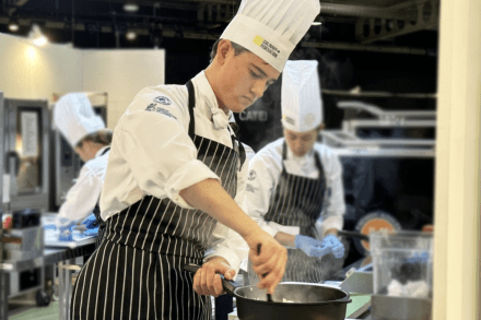 Ottis Crabbe wearing chef's hat and full cooking attire preparing a dish in a pot on a stove in an industrial kitchen during a competition.