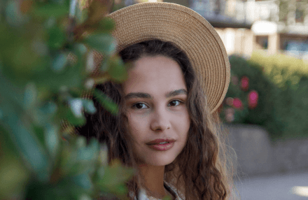 Anna Shynkarenko wearing a hat, half-hidden behind a bush and looking at the camera