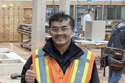 Ezat Haidary wering a reflective vests and safety goggles in the carpentry shop, giving a thumbs up and smiling at the camera.
