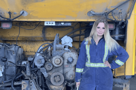 Madeline kozubal wearing mechanics coveralls standing in front of a school bus engine and smiling at the camera