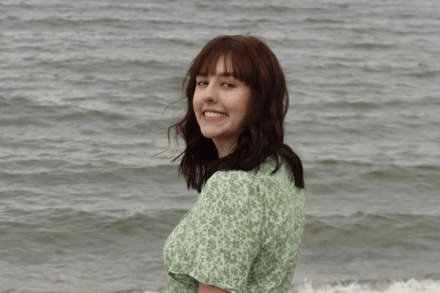 Paige Pierce standing in front of the ocean, wearing a green dress and turning back to smile at the camera