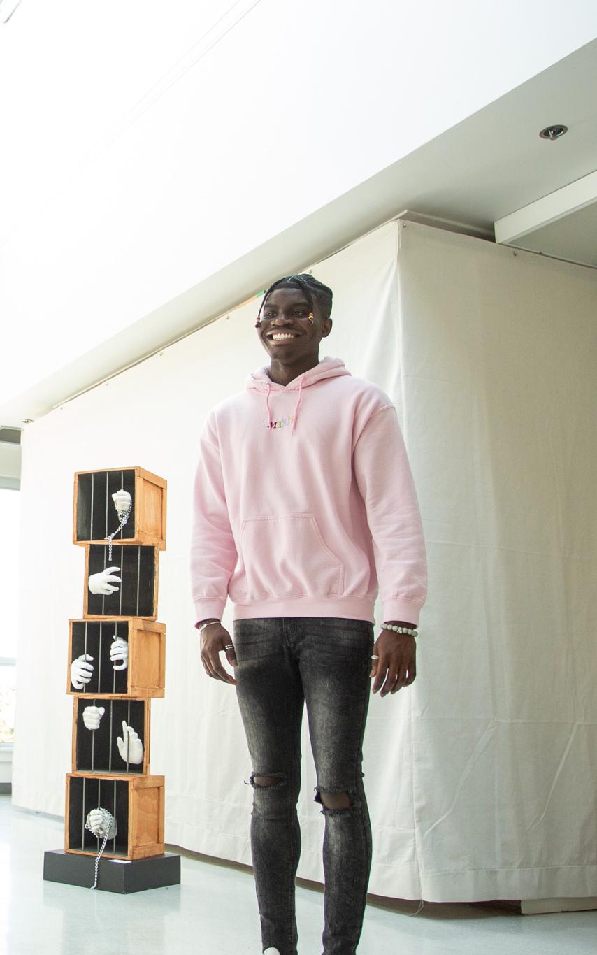 A smiling black man wearing a pink sweatshirt in an art gallery.